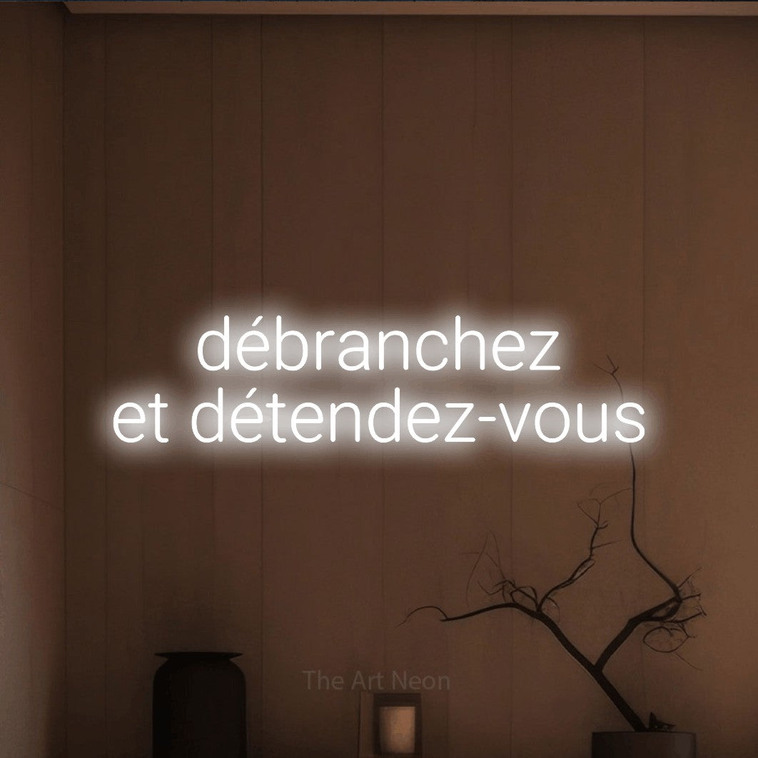 “Débranchez et détendez-vous” LED neon sign in cool white, with rounded sans-serif lettering across 2 lines, shown against a simple earth-toned interior.