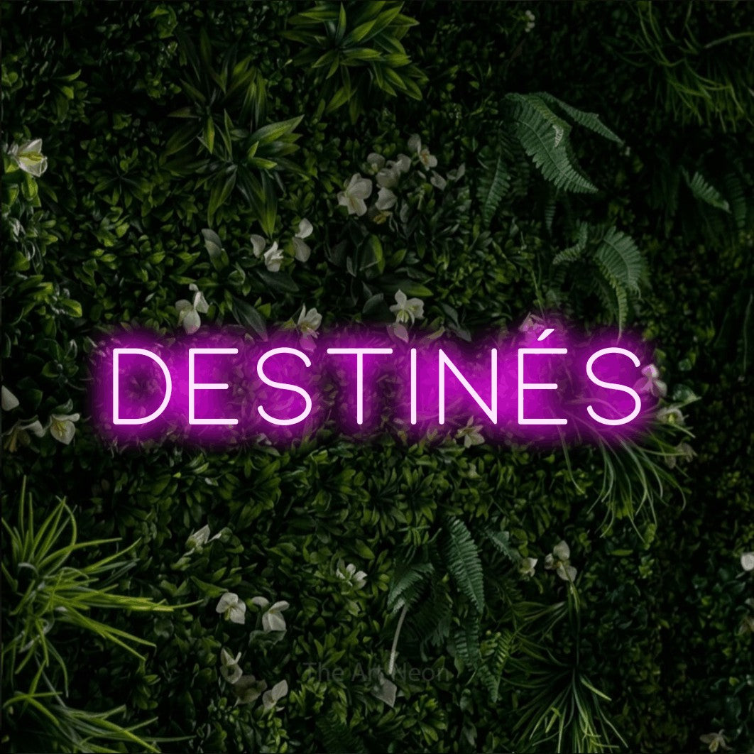 “Destinés” LED neon sign in pink, with thin, widely spaced lettering, shown against a dense green plant wall.
