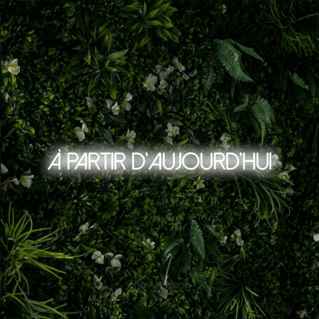 “À partir d'aujourd'hui” LED neon sign in cool white, with thin sans-serif lettering, shown against a dense green plant wall.