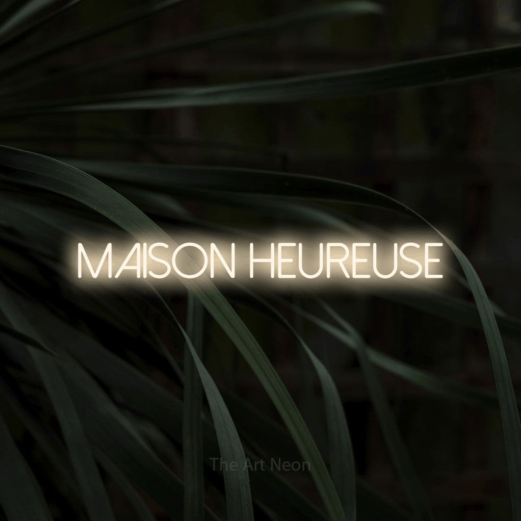 “Maison heureuse” LED neon sign in warm white, with thin sans-serif lettering, shown against a close-up of dark foliage.