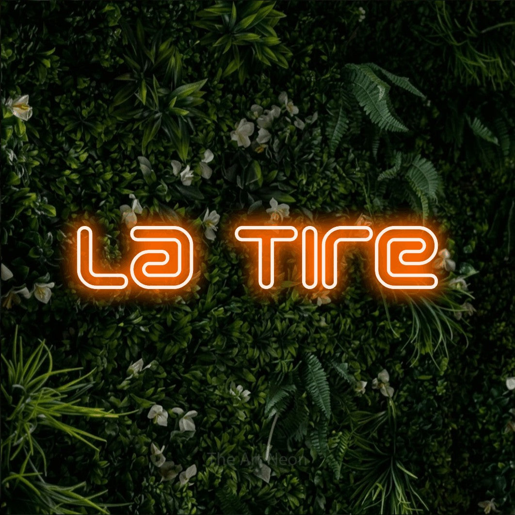 “La tire” LED neon sign in orange, with futuristic double-line lettering, shown against a dense green plant wall.