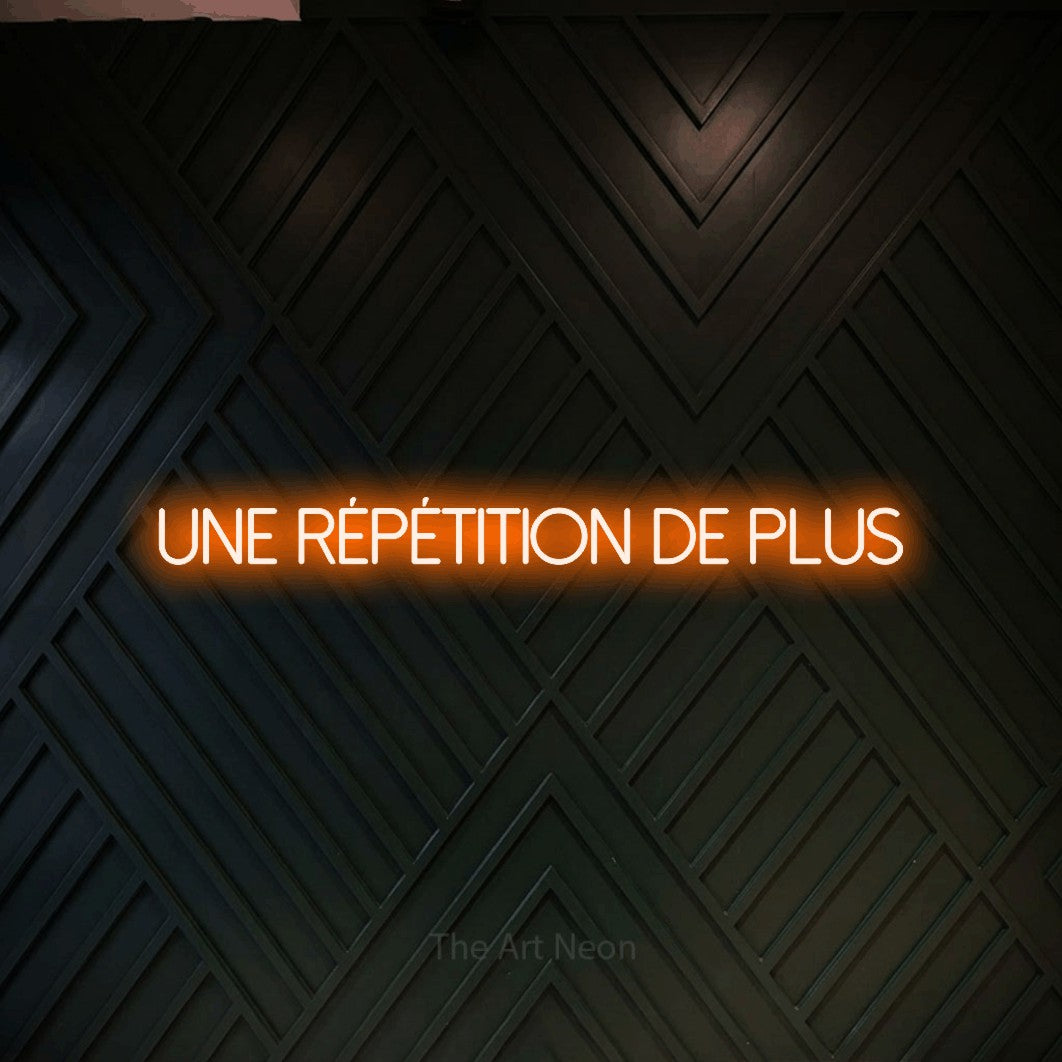 Décoration murale en néon LED « Une répétition de plus » en orange, en lettres fines sans empattement, installée sur un mur en bois noir à motif chevron, pour un espace d’entraînement.