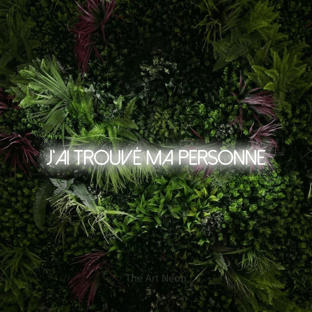 “J'ai trouvé ma personne” LED neon sign in cool white, with thin sans-serif lettering, shown against a plant wall with pink flowers.