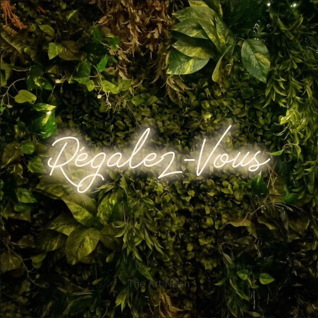 “Régalez-vous” LED neon sign in warm white, with cursive lettering, shown against bright green and yellow foliage.