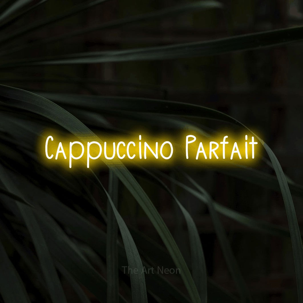 “Cappuccino parfait” LED neon sign in yellow, with narrow hand-drawn lettering, shown against a close-up of dark foliage.