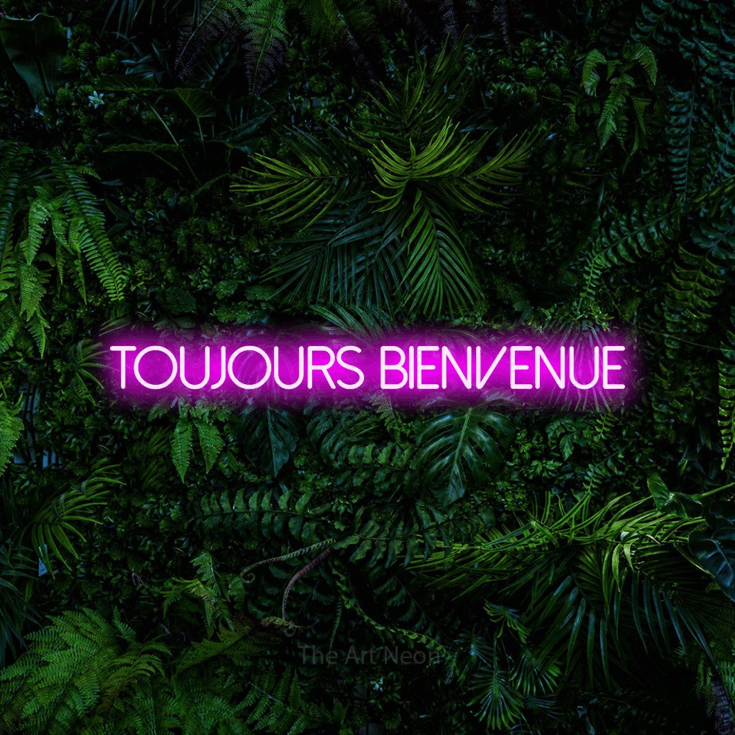 “Toujours bienvenue” LED neon sign in pink, with thin sans-serif lettering, shown against dense tropical foliage.