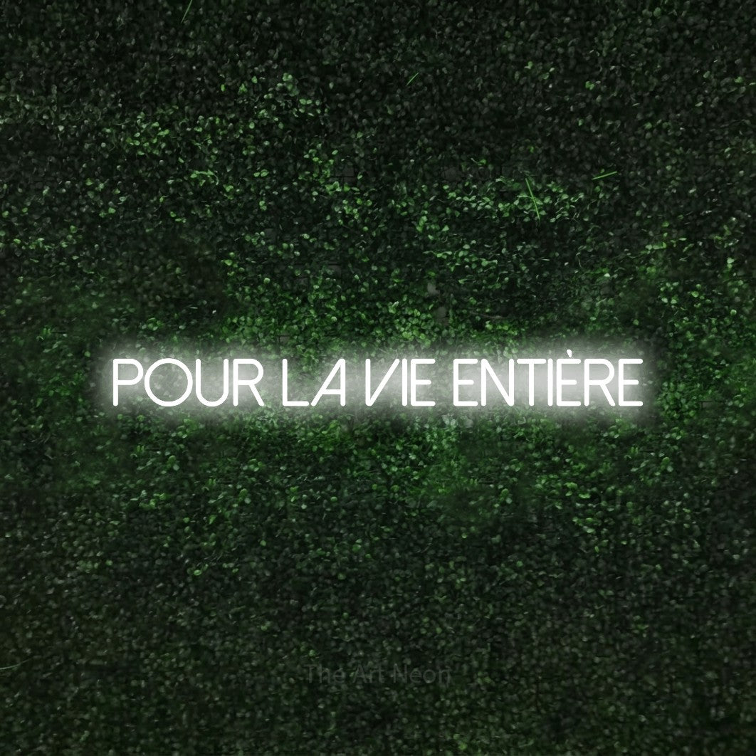 “Pour la vie entière” LED neon sign in cool white, with thin sans-serif lettering, shown against a textured green hedge.