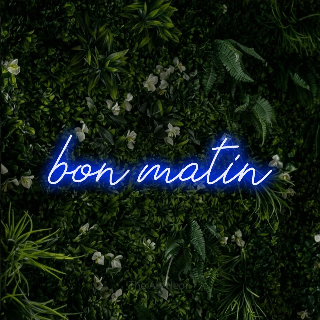 “Bon matin” LED neon sign in blue, with cursive lettering, shown against a dense green plant wall.