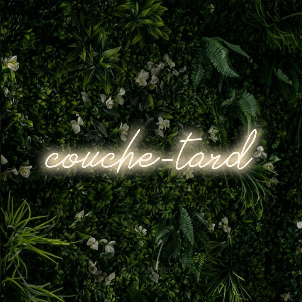 “Couche-tard” LED neon sign in warm white, with cursive lettering, shown against a dense green plant wall.