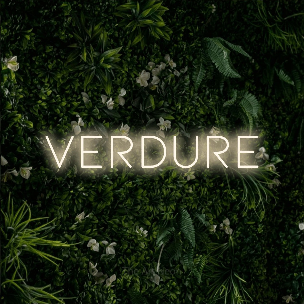 “Verdure” LED neon sign in warm white, with thin, widely spaced lettering, shown against a dense green plant wall.