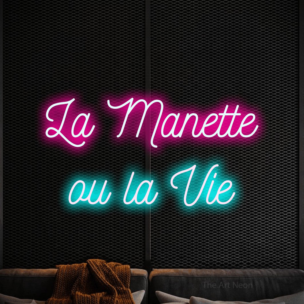 “La manette ou la vie” LED neon sign in hot pink on the upper line and ice blue on the lower line, with ornate cursive lettering across 2 lines, shown against a modern dark-toned seating area.
