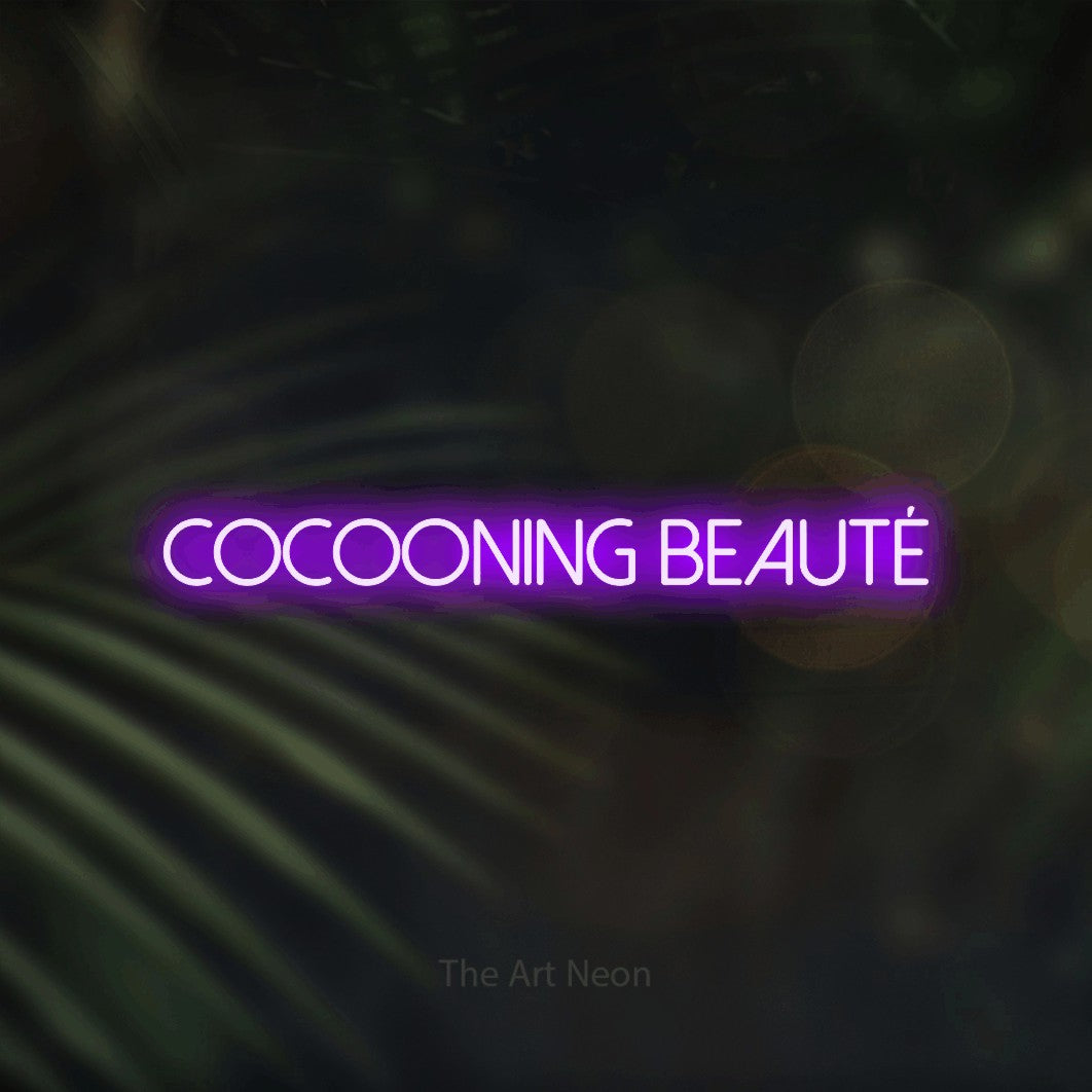 Composition en néon LED « Cocooning beauté » en violet, en lettres fines sans empattement, mise en valeur sur un fond flou de palmiers et de lumières bokeh, pour un salon de beauté.