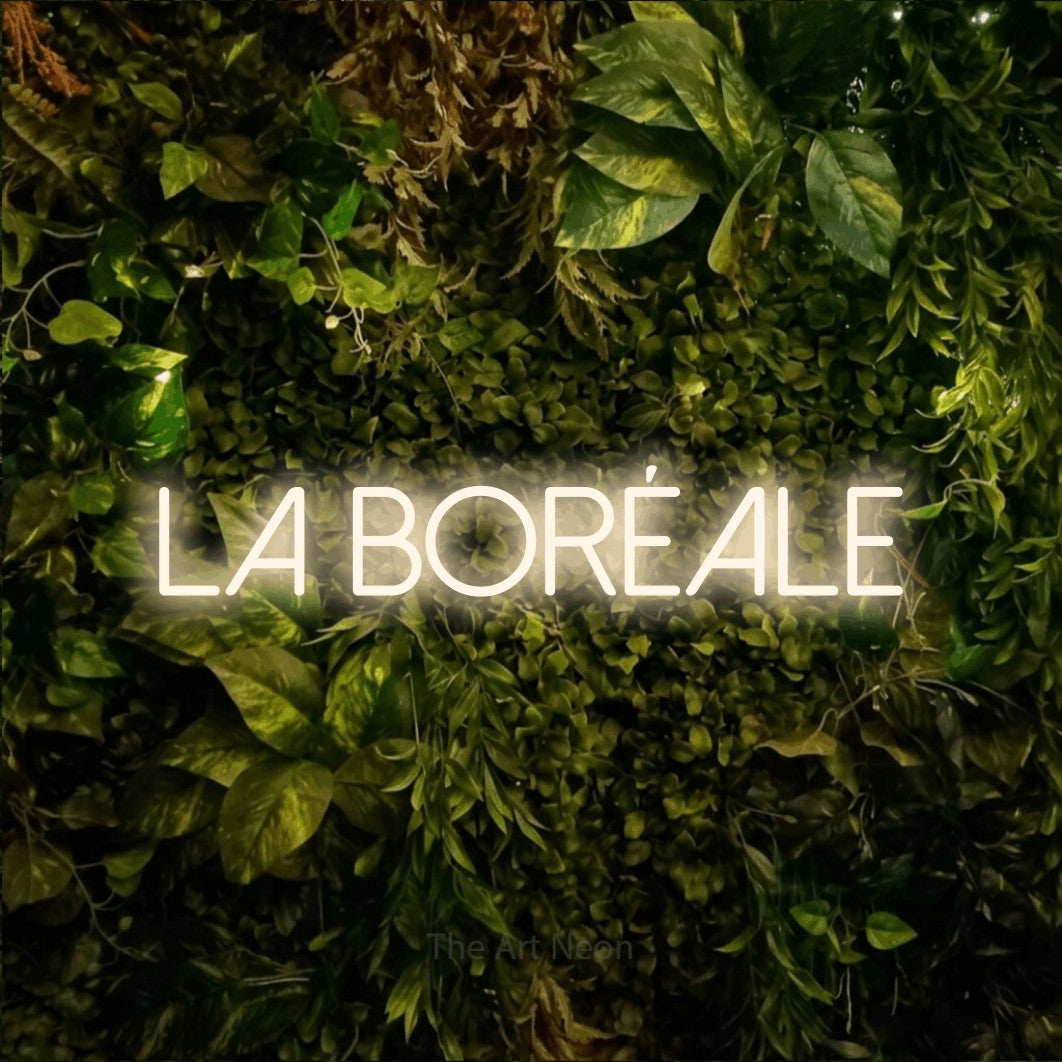 “La boréale” LED neon sign in warm white, with thin sans-serif lettering, shown against bright green and yellow foliage.