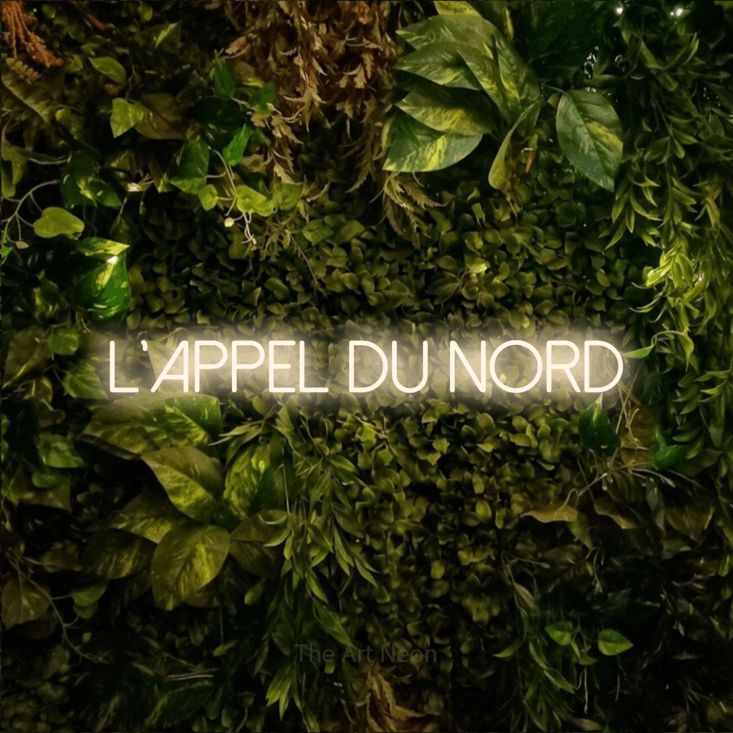 Lettrage en néon LED « L'appel du nord » en blanc chaud, en lettres fines sans empattement, présenté sur un feuillage vert et jaune vif, pour un décor de plein air canadien.