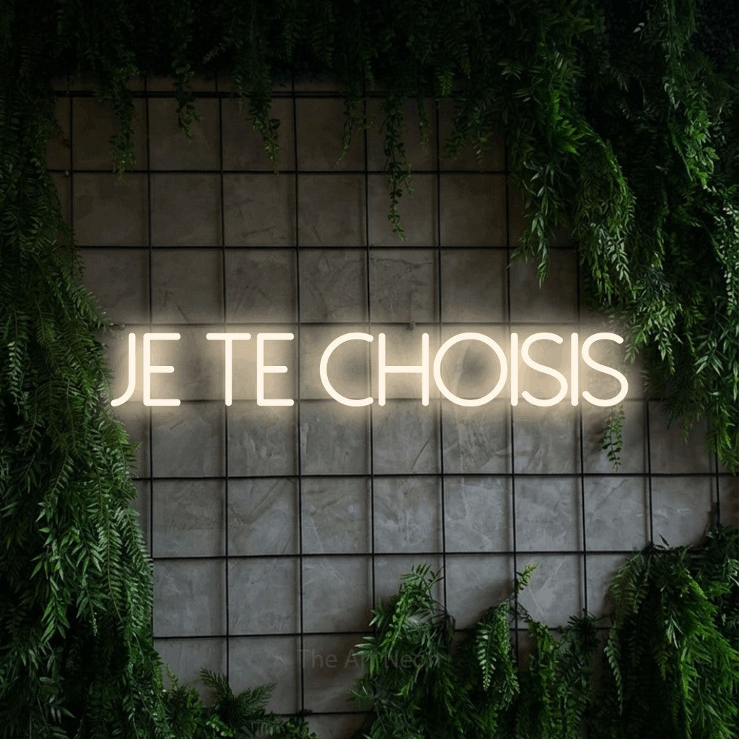 “Je te choisis” LED neon sign in warm white, with thin sans-serif lettering, shown against an ivy-covered concrete grid.