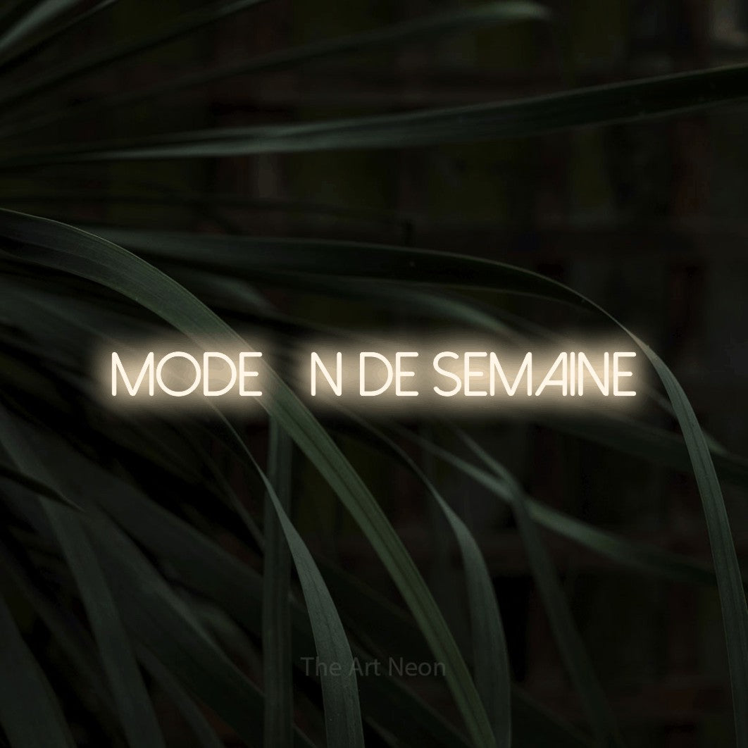 “Mode fin de semaine” LED neon sign in warm white, with thin sans-serif lettering, shown against a close-up of dark foliage.