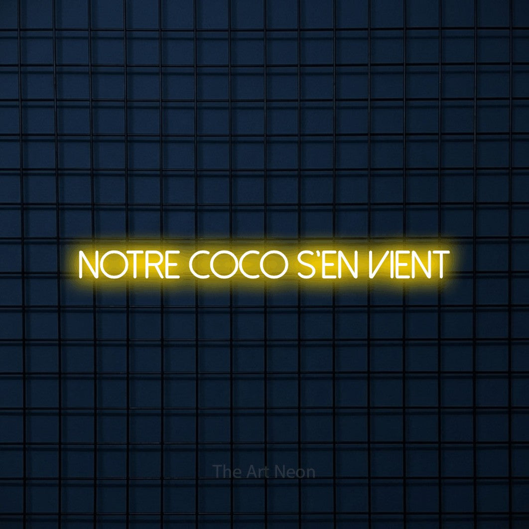 Aperçu d’une enseigne néon LED « Notre coco s'en vient » en jaune, en lettres fines sans empattement, photographiée sur une grille bleu foncé, pour une fête prénatale.