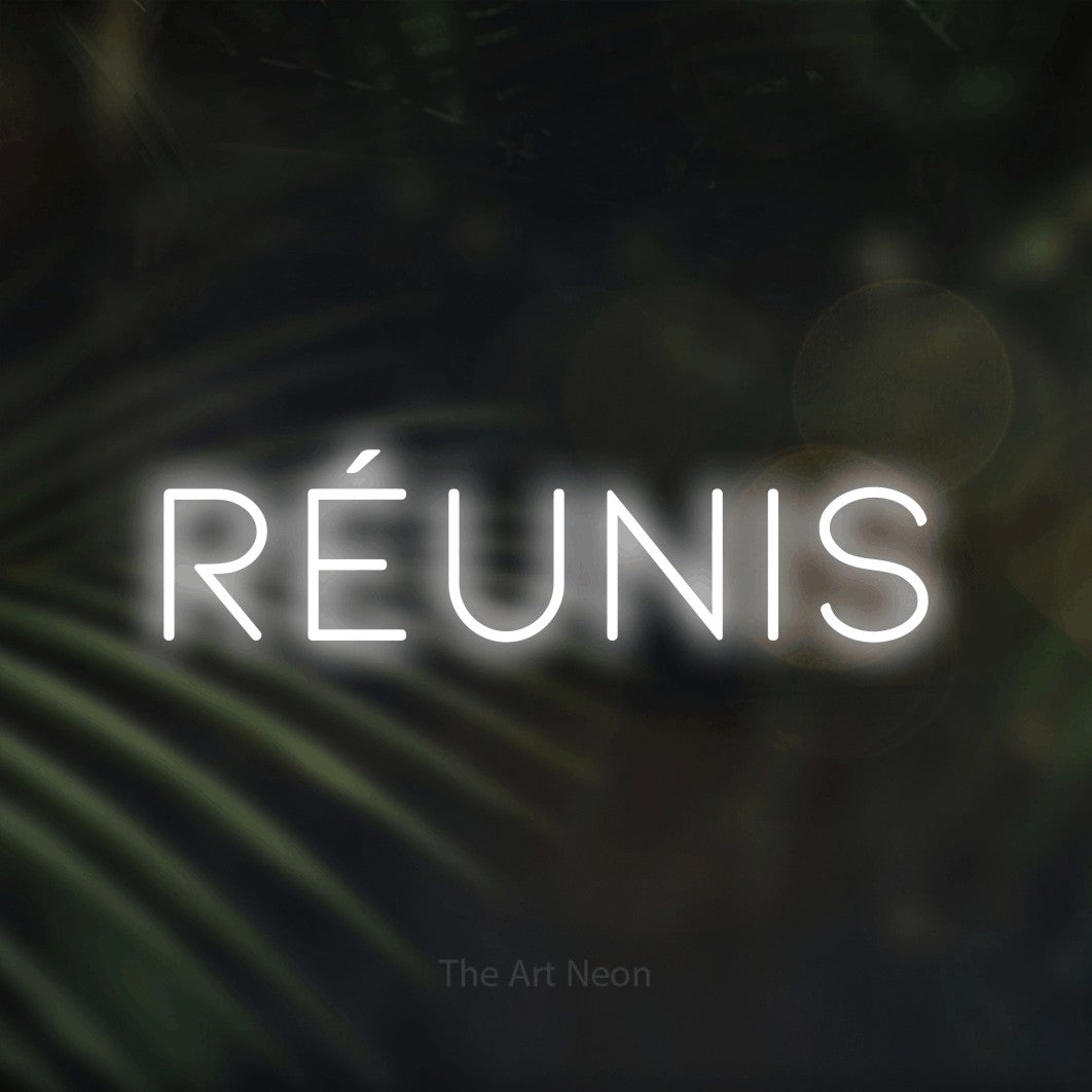 “Réunis” LED neon sign in cool white, with thin, widely spaced lettering, shown against a blurred palm and bokeh-light backdrop.