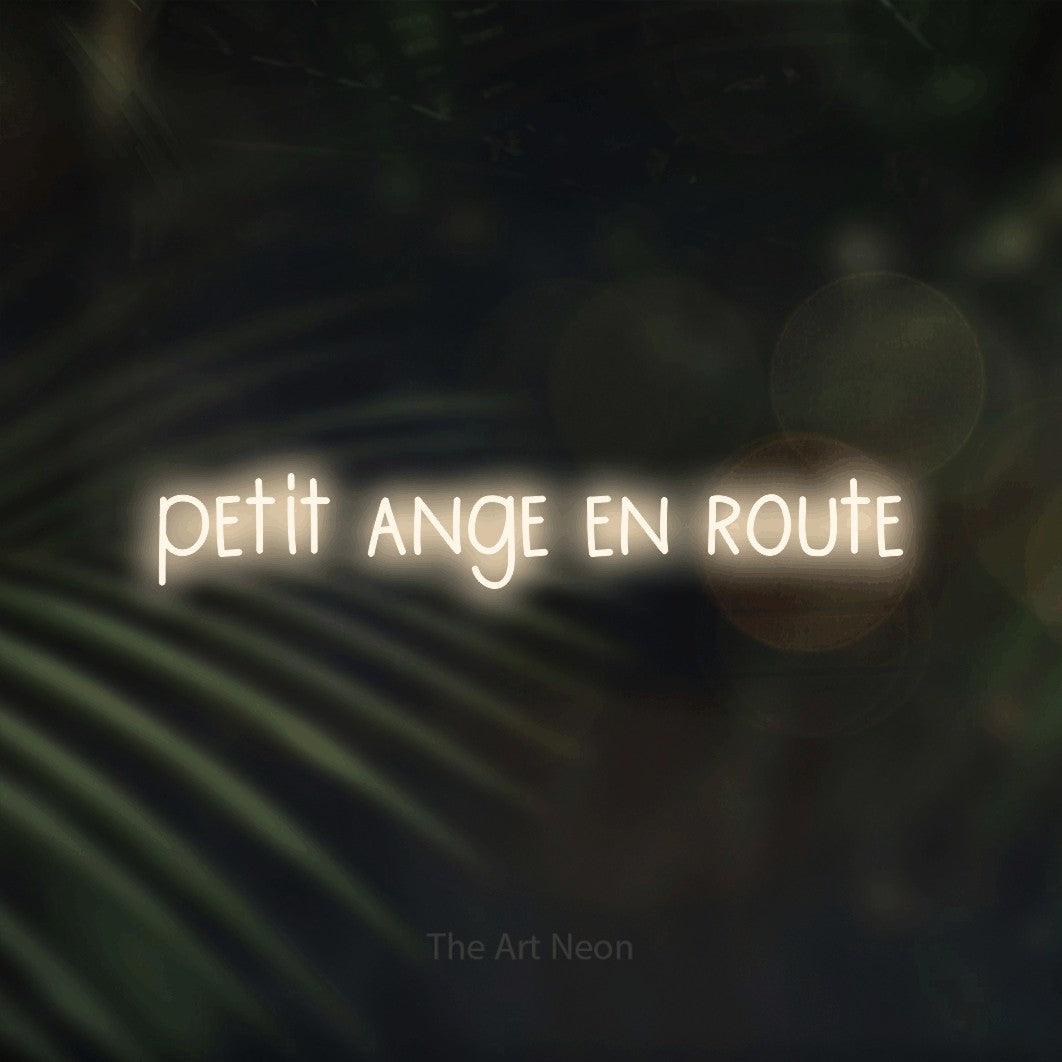 “Petit ange en route” LED neon sign in warm white, with narrow hand-drawn lettering, shown against a blurred palm and bokeh-light backdrop.