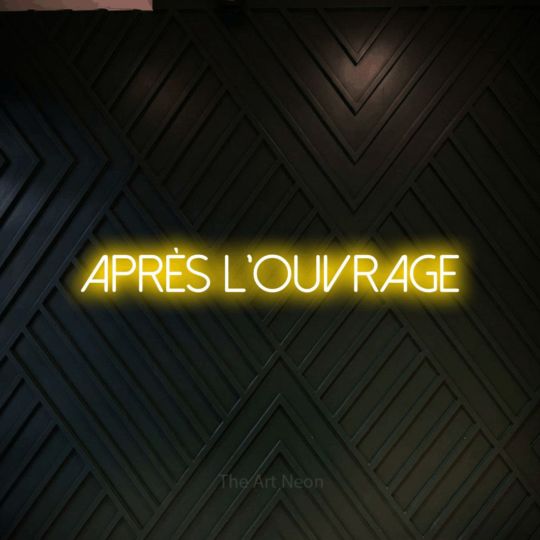 “Après l'ouvrage” LED neon sign in yellow, with thin sans-serif lettering, shown against a black chevron-patterned wood wall.