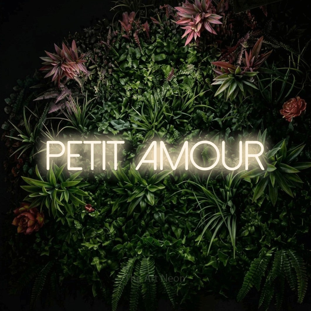 “Petit amour” LED neon sign in warm white, with thin sans-serif lettering, shown against a flowering plant wall.