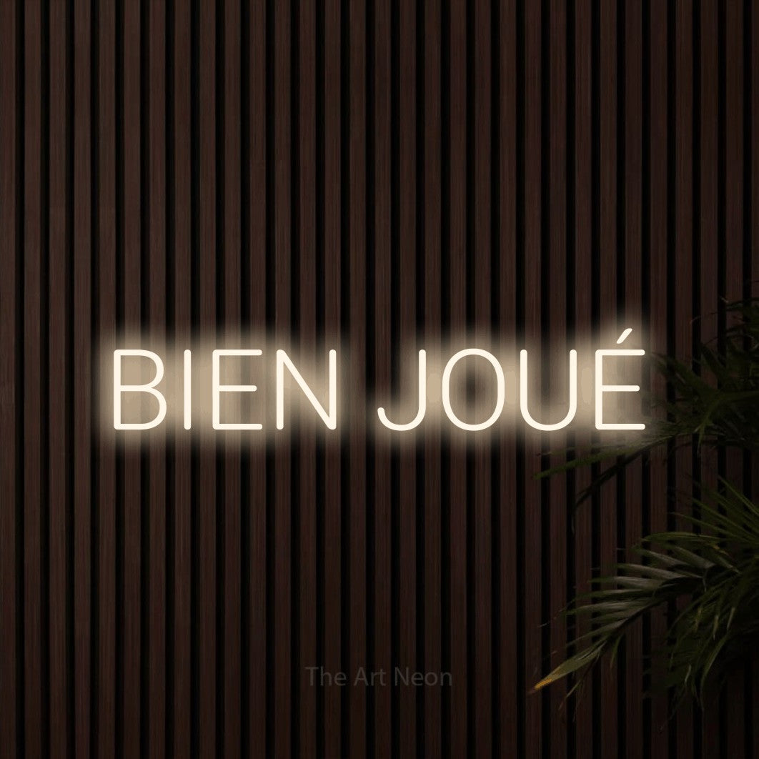 “Bien joué” LED neon sign in warm white, with rounded sans-serif lettering, shown against dark wooden wall slats.
