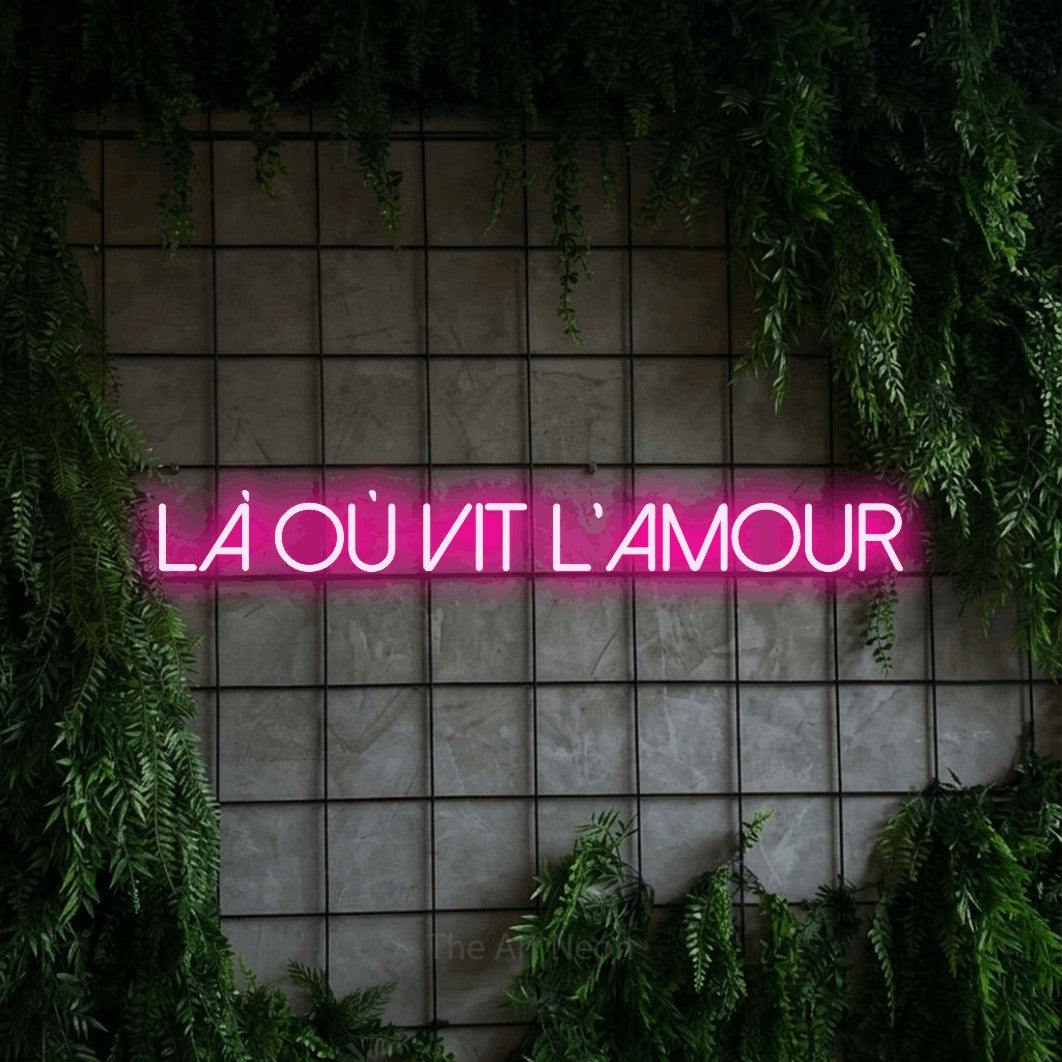 “Là où vit l'amour” LED neon sign in hot pink, with thin sans-serif lettering, shown against an ivy-covered concrete grid.