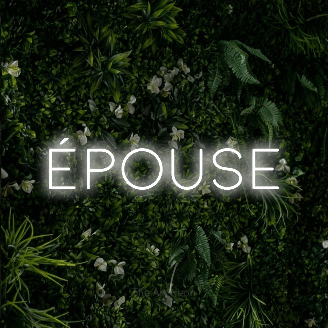 “Épouse” LED neon sign in cool white, with thin, widely spaced lettering, shown against a dense green plant wall.