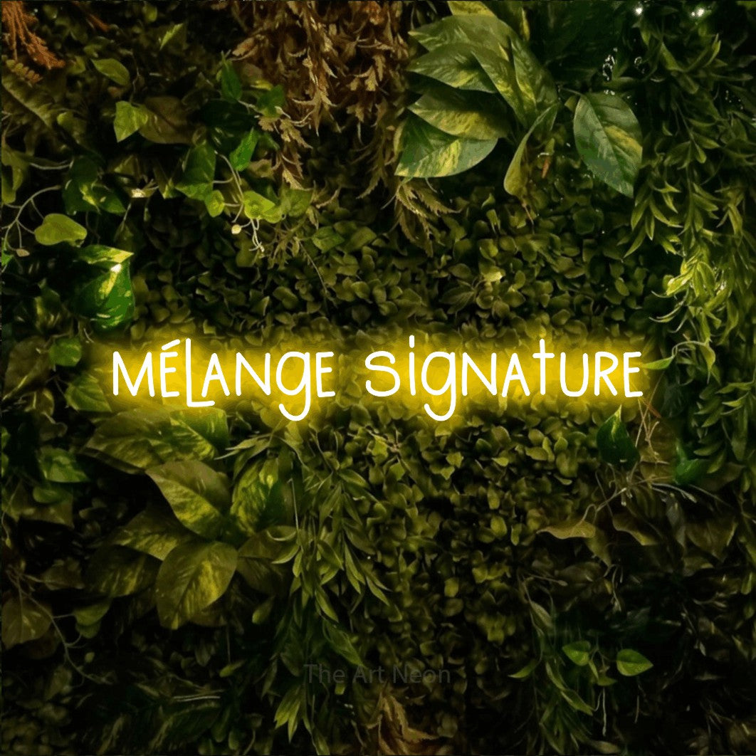 “Mélange signature” LED neon sign in yellow, with narrow hand-drawn lettering, shown against bright green and yellow foliage.
