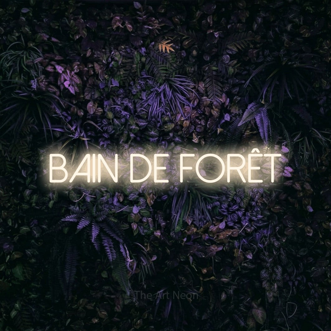 “Bain de forêt” LED neon sign in warm white, with thin sans-serif lettering, shown against a dark wall with purple flowers.