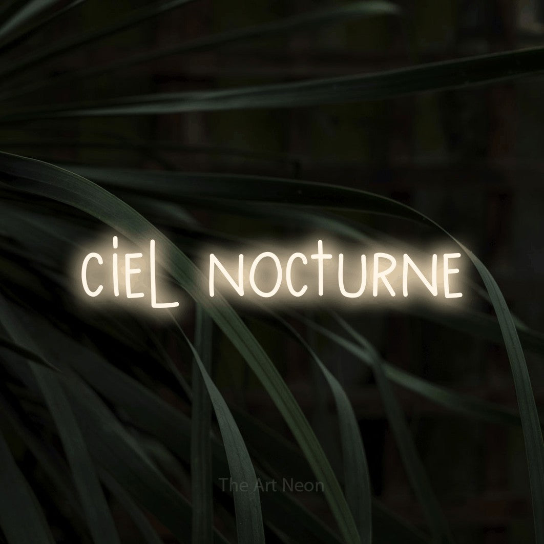 “Ciel nocturne” LED neon sign in warm white, with narrow hand-drawn lettering, shown against a close-up of dark foliage.
