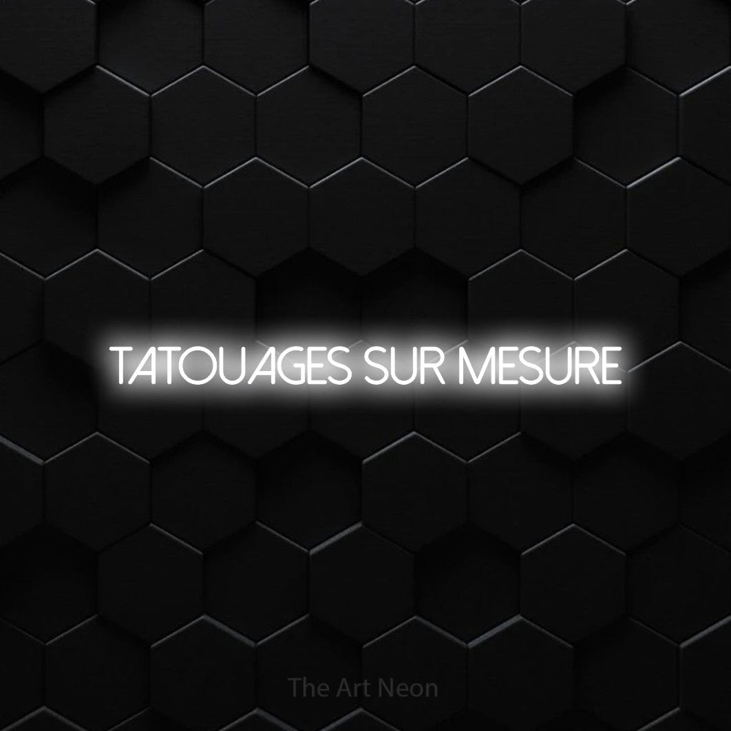 “Tatouages sur mesure” LED neon sign in cool white, with thin sans-serif lettering, shown against black hexagonal wall tiles.