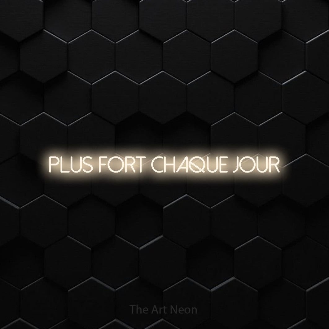 “Plus fort chaque jour” LED neon sign in warm white, with thin sans-serif lettering, shown against black hexagonal wall tiles.