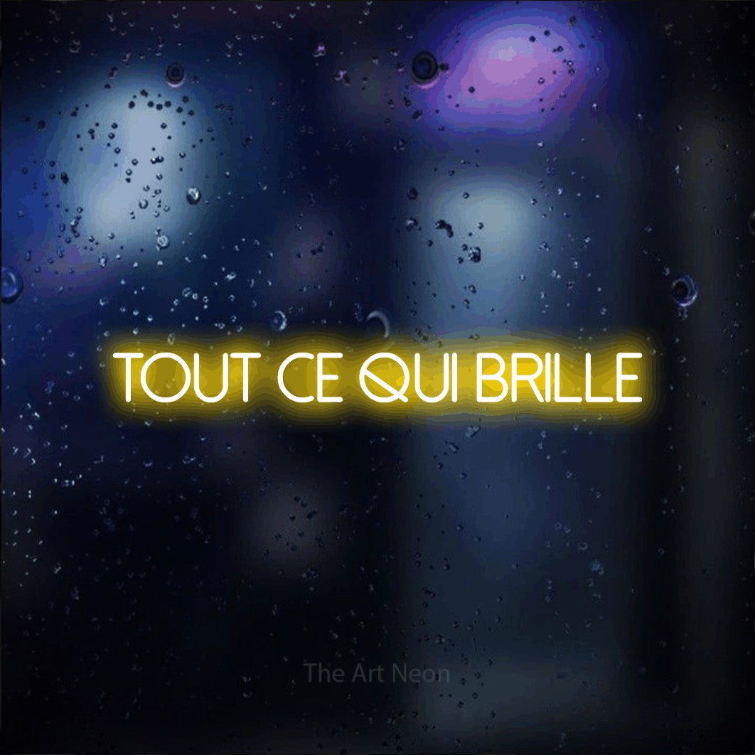 “Tout ce qui brille” LED neon sign in yellow, with thin sans-serif lettering, shown against glass with blue and purple reflections.