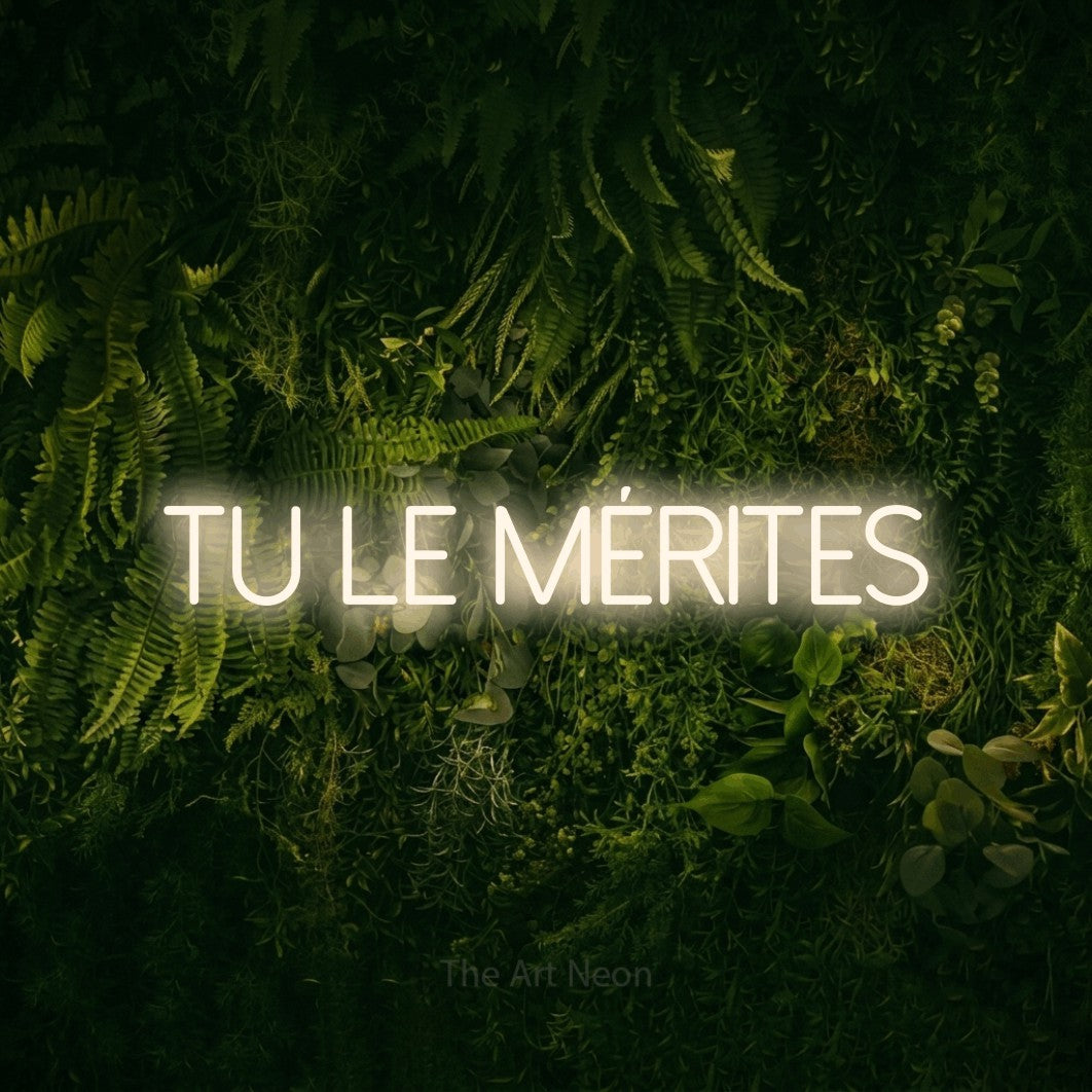 “Tu le mérites” LED neon sign in warm white, with thin sans-serif lettering, shown against a dark moss and forest backdrop.