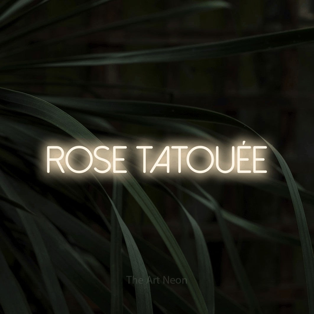 “Rose tatouée” LED neon sign in warm white, with thin sans-serif lettering, shown against a close-up of dark foliage.