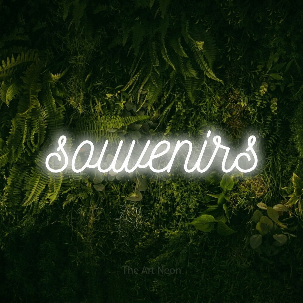 “Souvenirs” LED neon sign in cool white, with ornate cursive lettering, shown against a dark moss and forest backdrop.