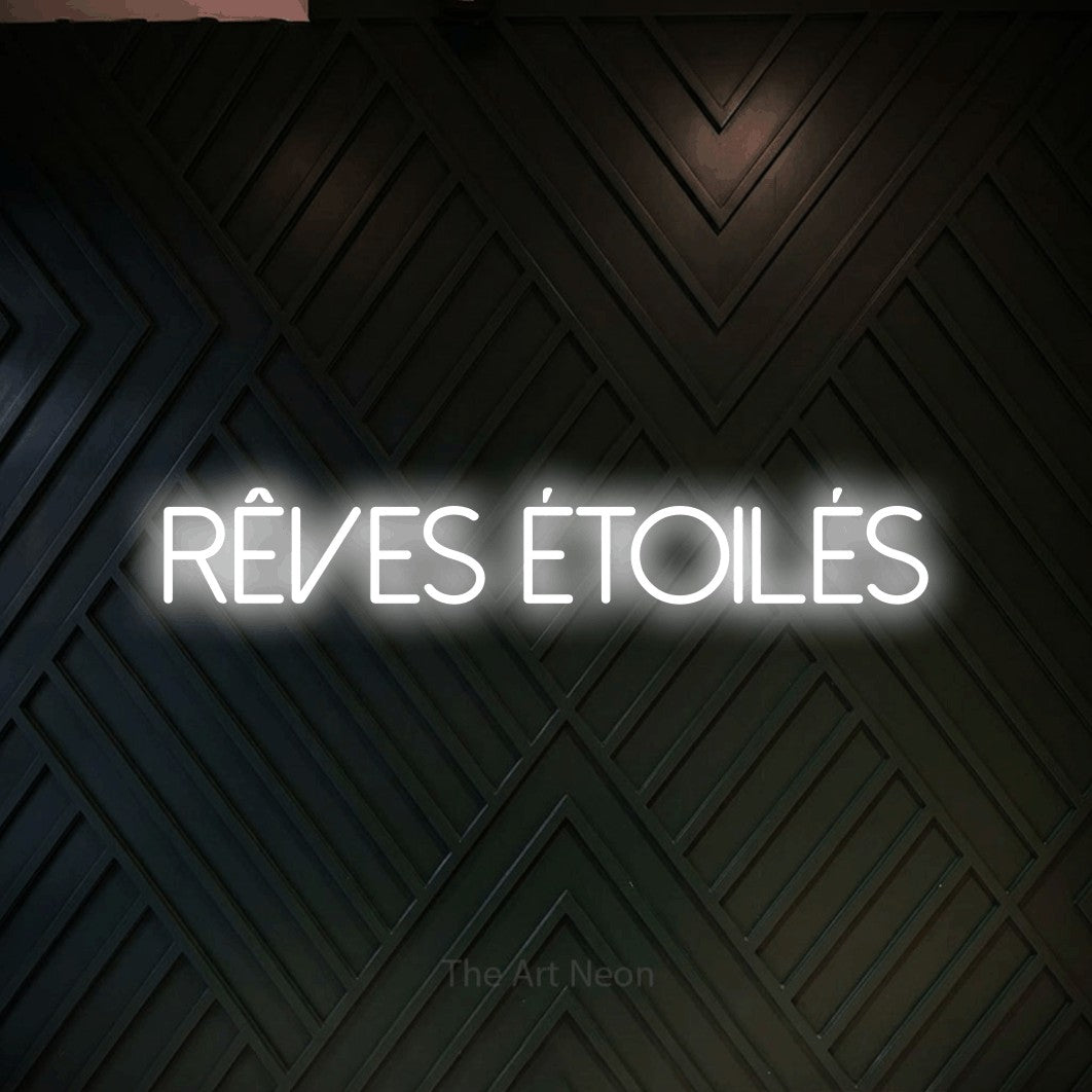 “Rêves étoilés” LED neon sign in cool white, with thin sans-serif lettering, shown against a black chevron-patterned wood wall.