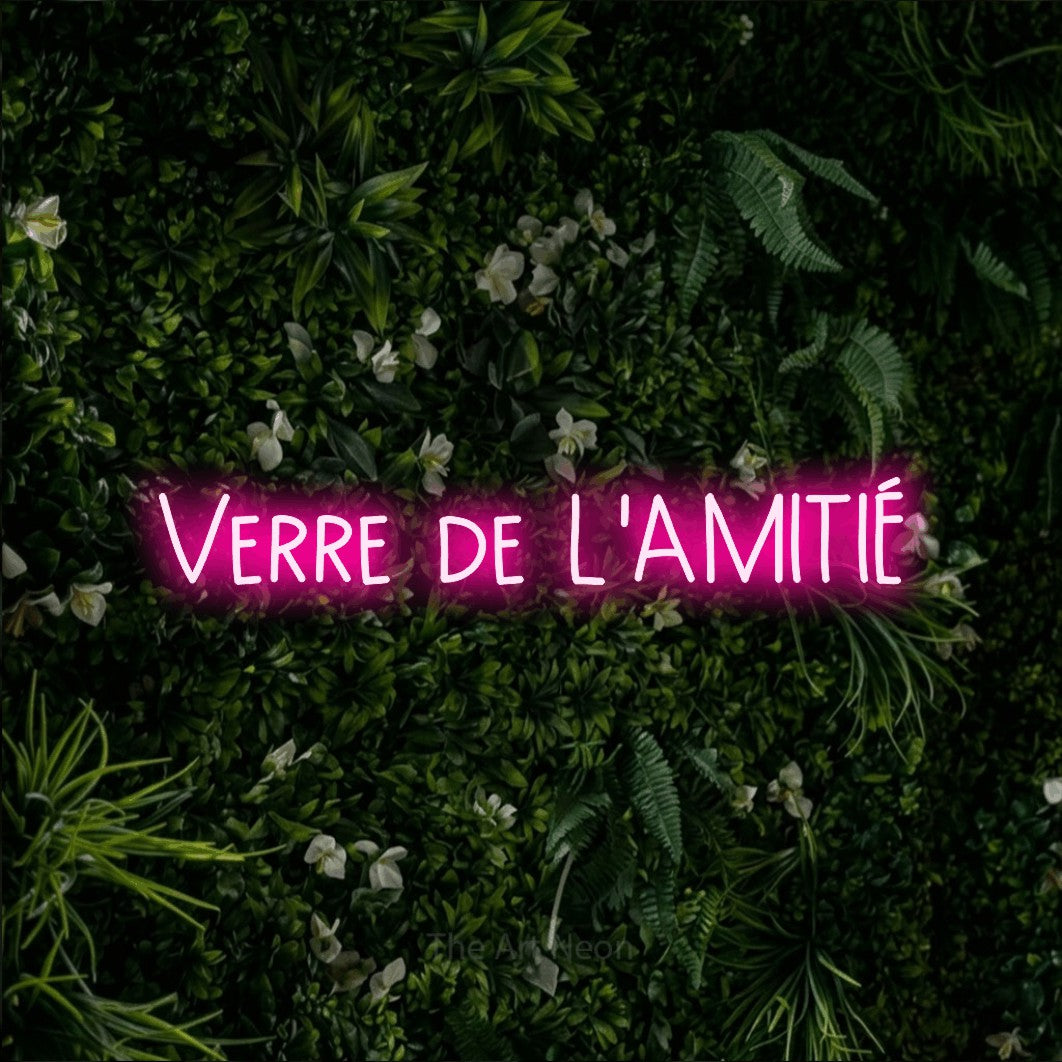 “Verre de l'amitié” LED neon sign in hot pink, with narrow hand-drawn lettering, shown against a dense green plant wall.