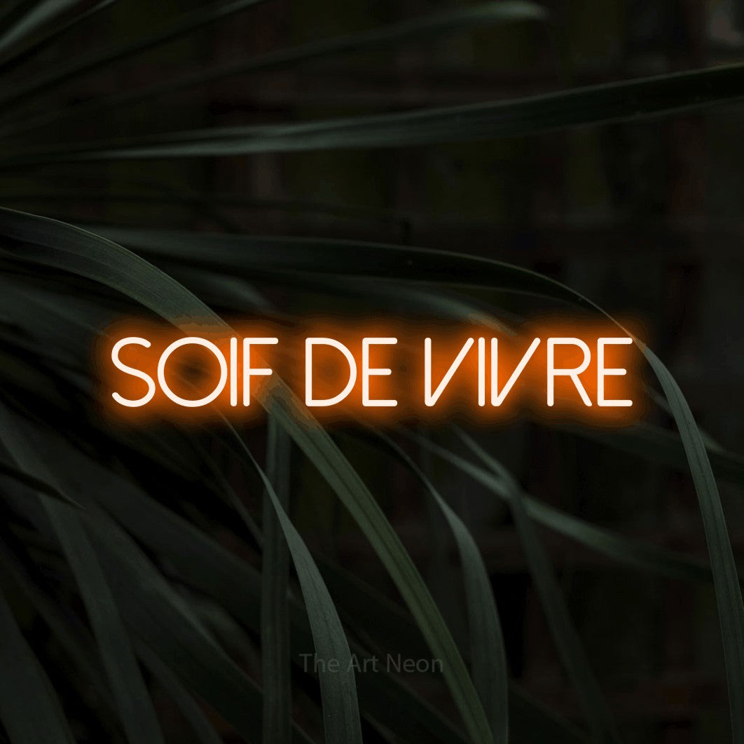 “Soif de vivre” LED neon sign in orange, with thin sans-serif lettering, shown against a close-up of dark foliage.