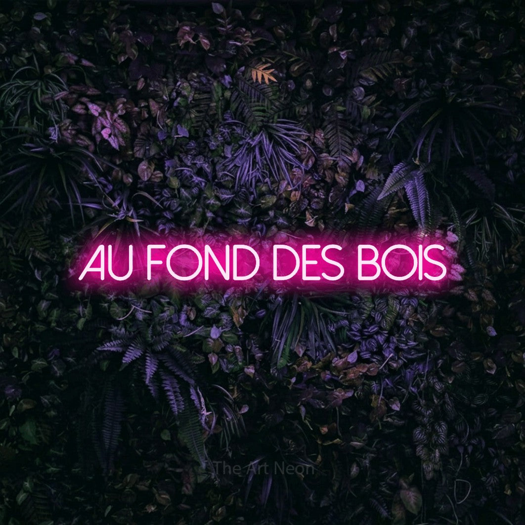 “Au fond des bois” LED neon sign in hot pink, with thin sans-serif lettering, shown against a dark wall with purple flowers.