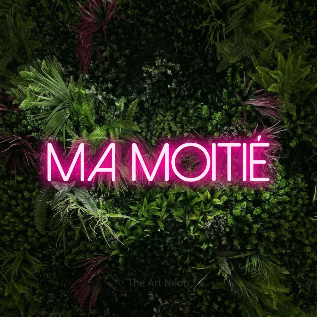 “Ma moitié” LED neon sign in hot pink, with thin sans-serif lettering, shown against a plant wall with pink flowers.