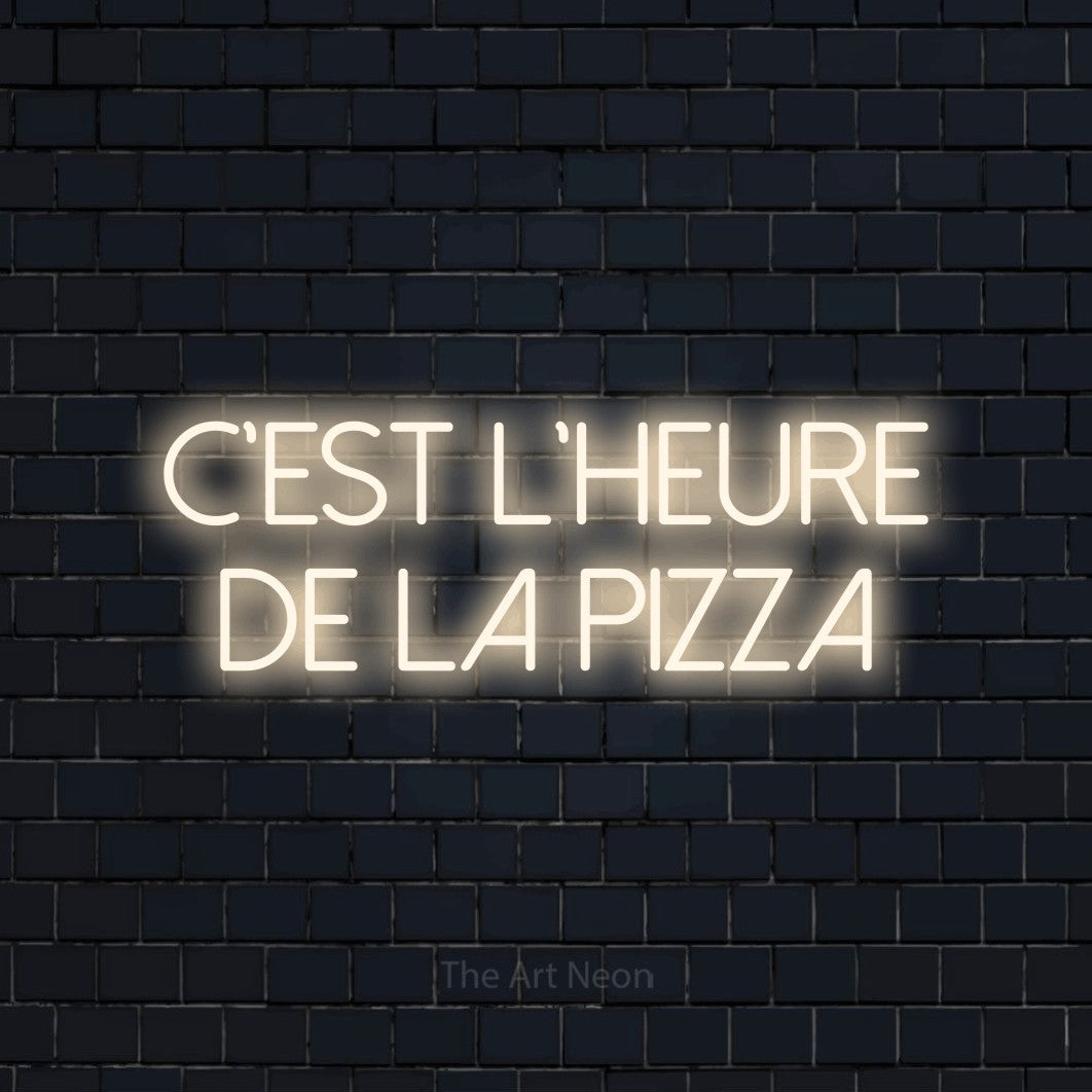 “C'est l'heure de la pizza” LED neon sign in warm white, with thin sans-serif lettering across 2 lines, shown against a dark navy brick wall.