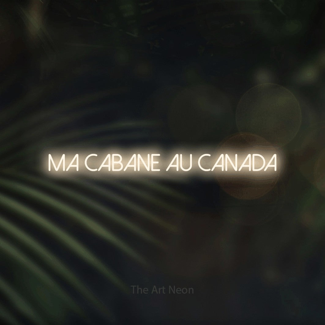 “Ma cabane au Canada” LED neon sign in warm white, with thin sans-serif lettering, shown against a blurred palm and bokeh-light backdrop.