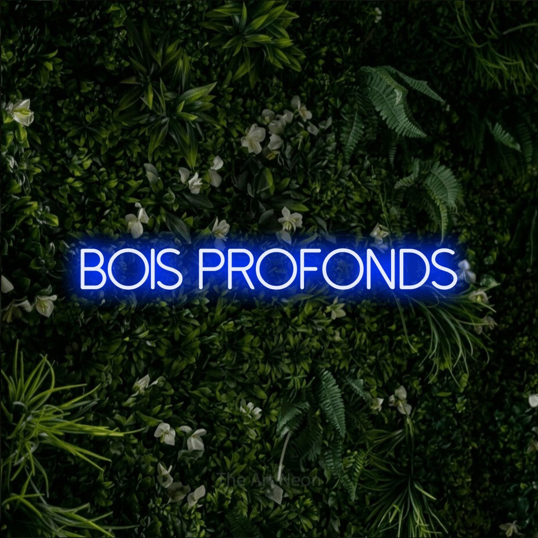 “Bois profonds” LED neon sign in blue, with thin sans-serif lettering, shown against a dense green plant wall.
