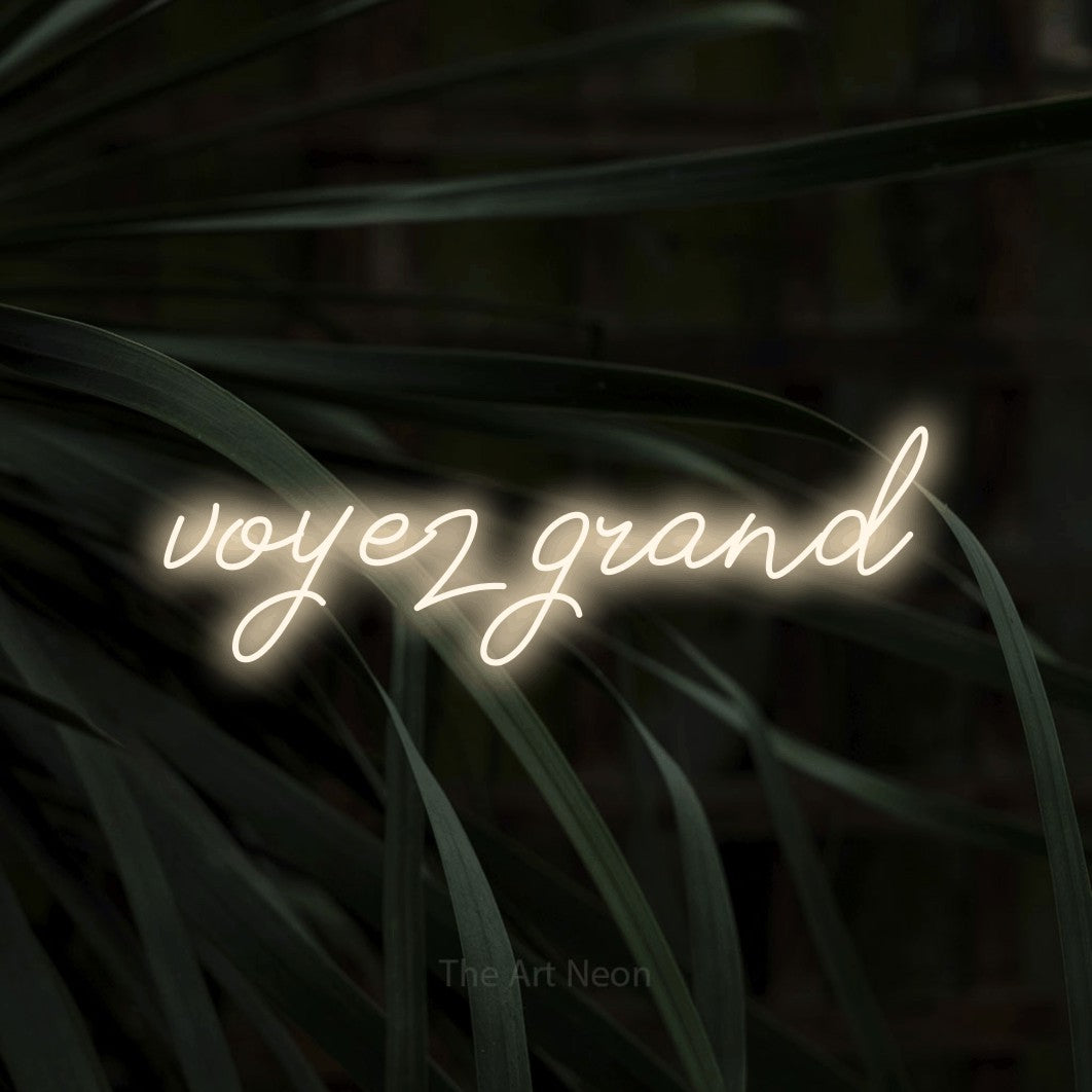“Voyez grand” LED neon sign in warm white, with cursive lettering, shown against a close-up of dark foliage.