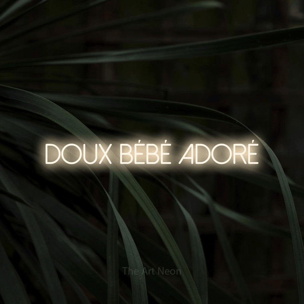 Décoration murale en néon LED « Doux bébé adoré » en blanc chaud, en lettres fines sans empattement, installée sur un feuillage sombre en gros plan, pour une fête prénatale.