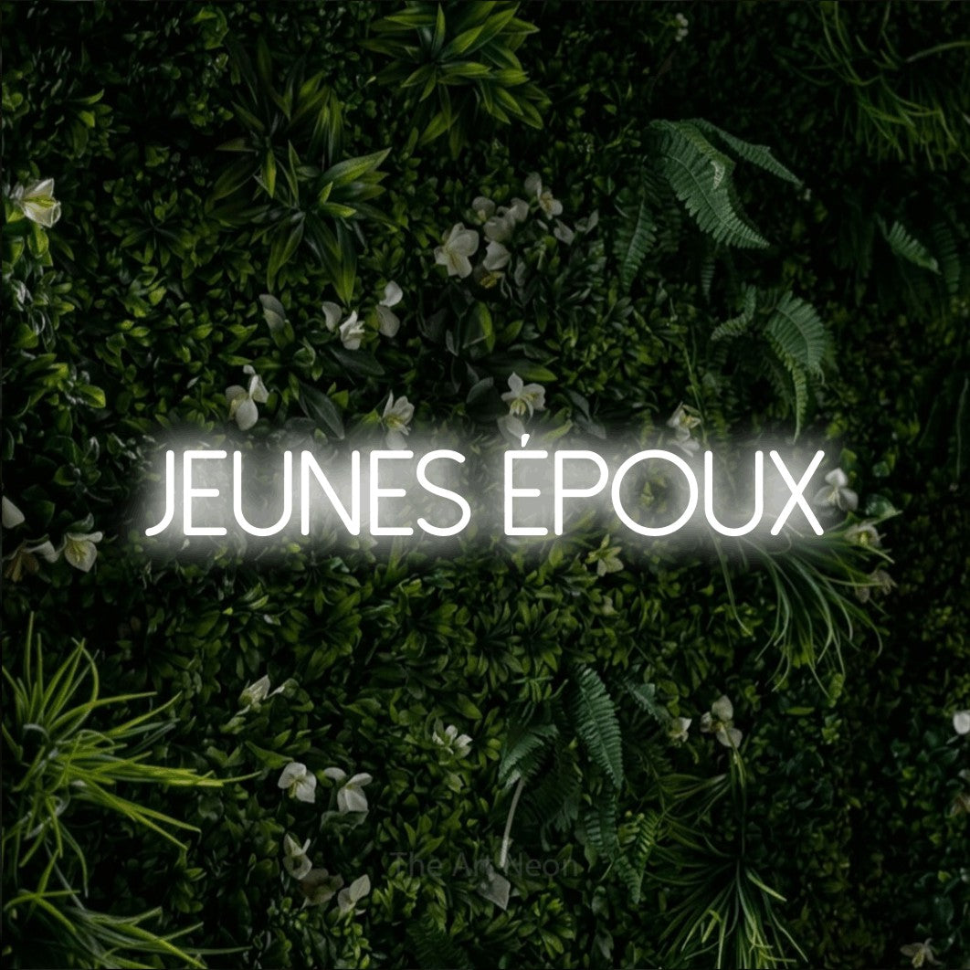 “Jeunes époux” LED neon sign in cool white, with thin sans-serif lettering, shown against a dense green plant wall.