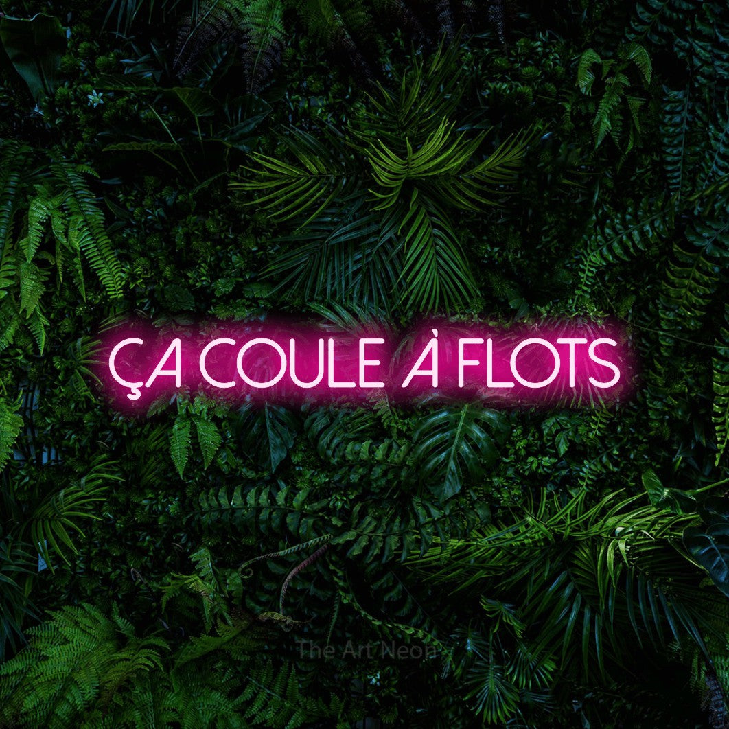 Composition en néon LED « Ça coule à flots » en rose vif, en lettres fines sans empattement, mise en valeur devant un feuillage tropical dense, pour un bar ou un pub.