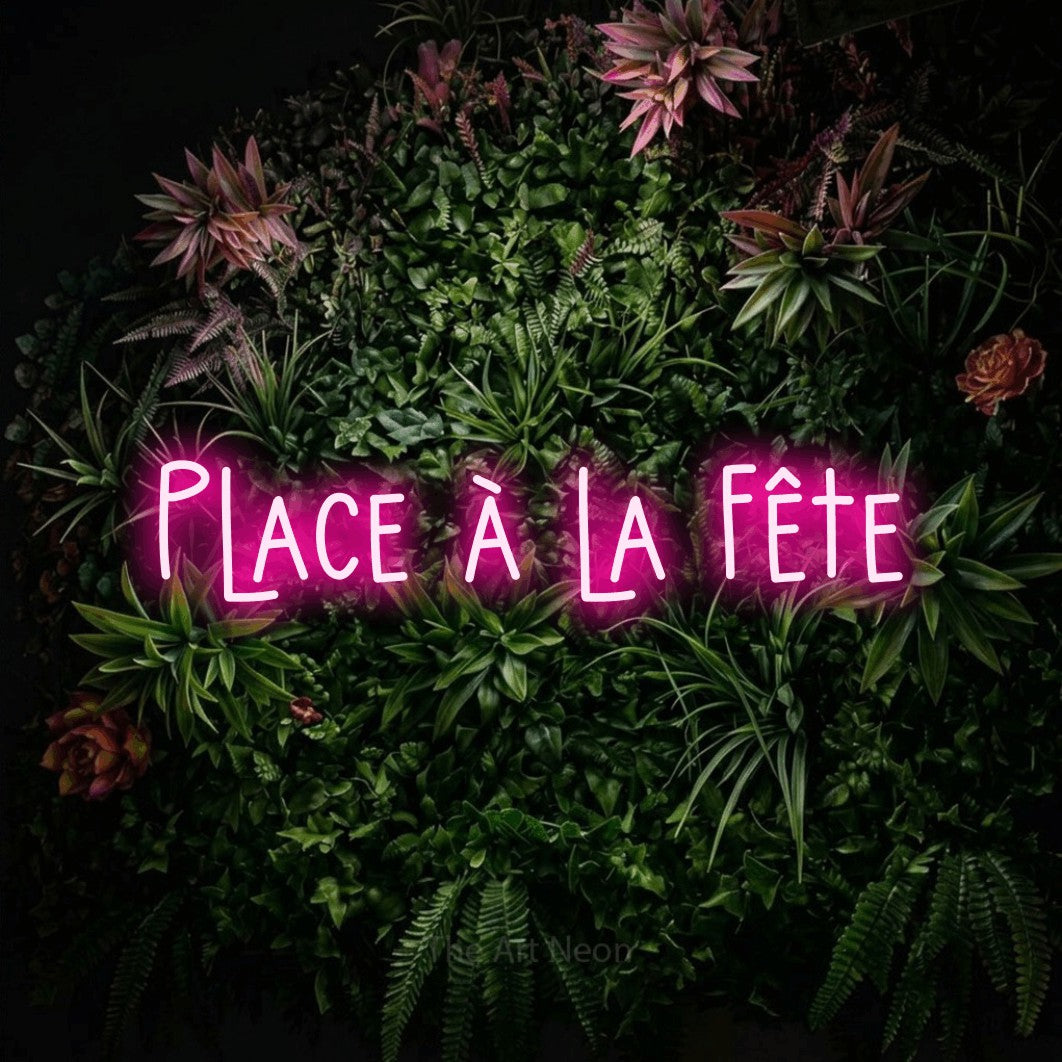 “Place à la fête” LED neon sign in hot pink, with narrow hand-drawn lettering, shown against a flowering plant wall.